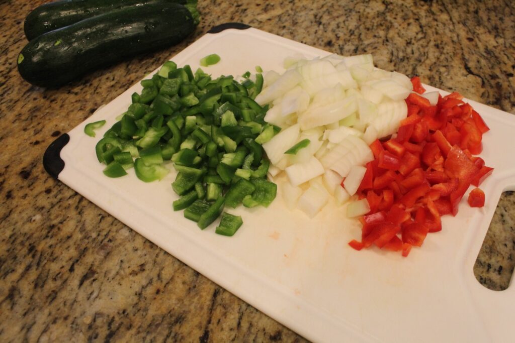 Diced bell peppers  and onions on a cutting board with zucchinis in the background.