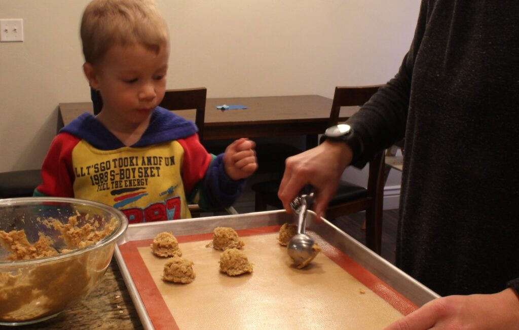 Boy helping scoop peanut butter oatmeal cookies into cookie sheet.