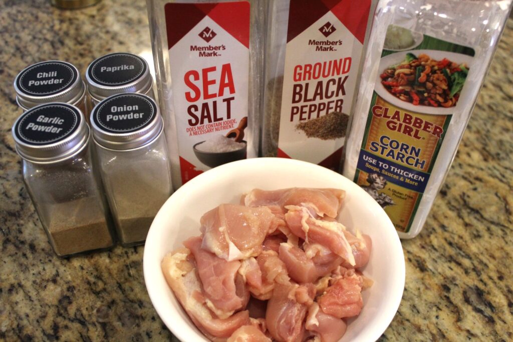 Seasonings and cornstarch next to a bowl of diced chicken.