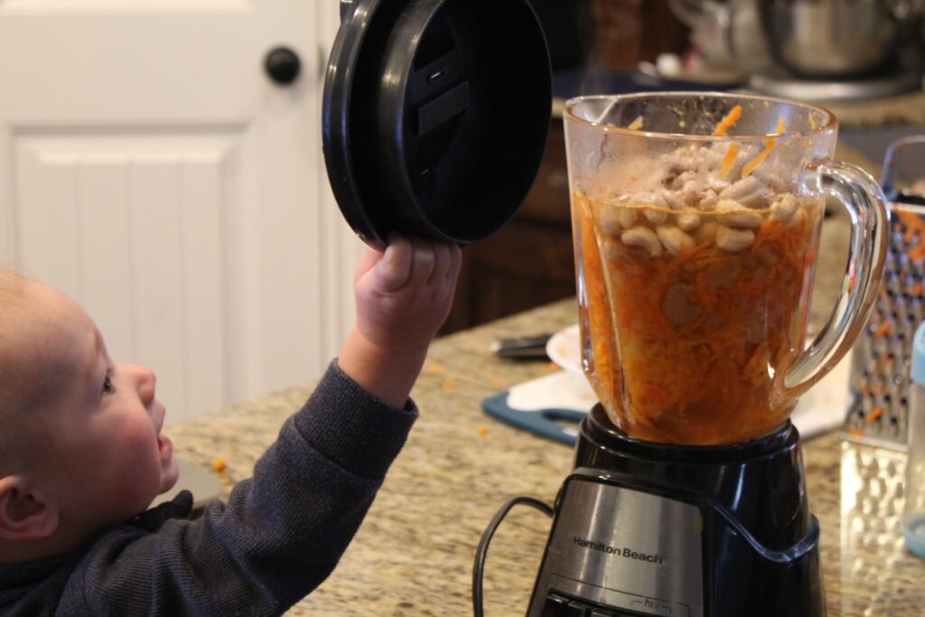 Small boy putting the lid on a blender full of boiled vegetables and cashews.