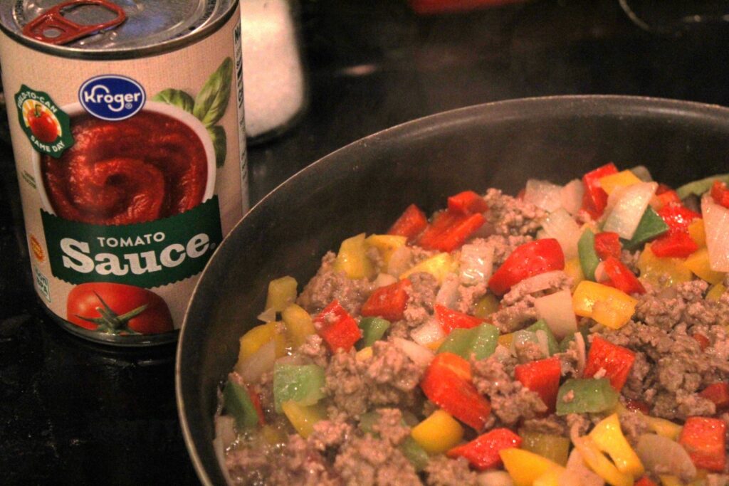 Ground beef, peppers and onion cooking in a pan beside a can of tomato sauce.