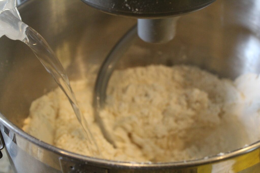 pouring a water mixture into a stand mixer full of a flour mixture. 