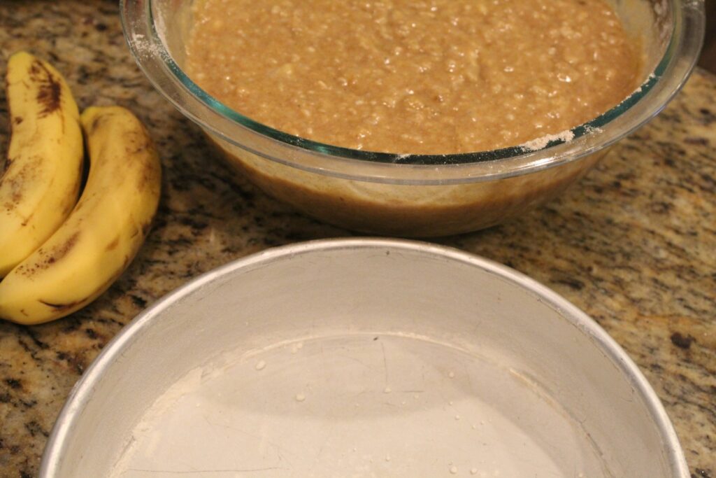 Bananas next to banana oat cake batter, and a greased and floured cake pan.