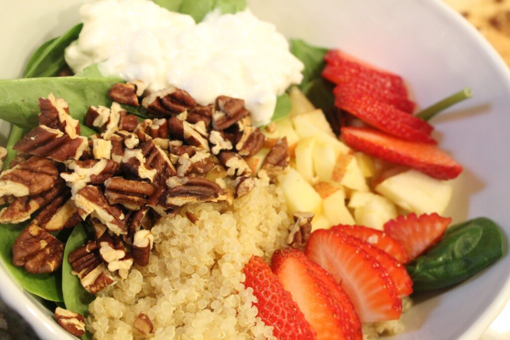 A bowl of spinach topped with strawberries, quinoa, pecans, apples, and cottage cheese.