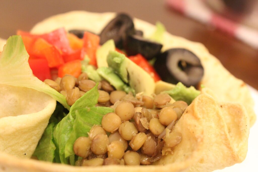 a taco salad bowl topped with lentils, avocado, tomatoes, and olives. 