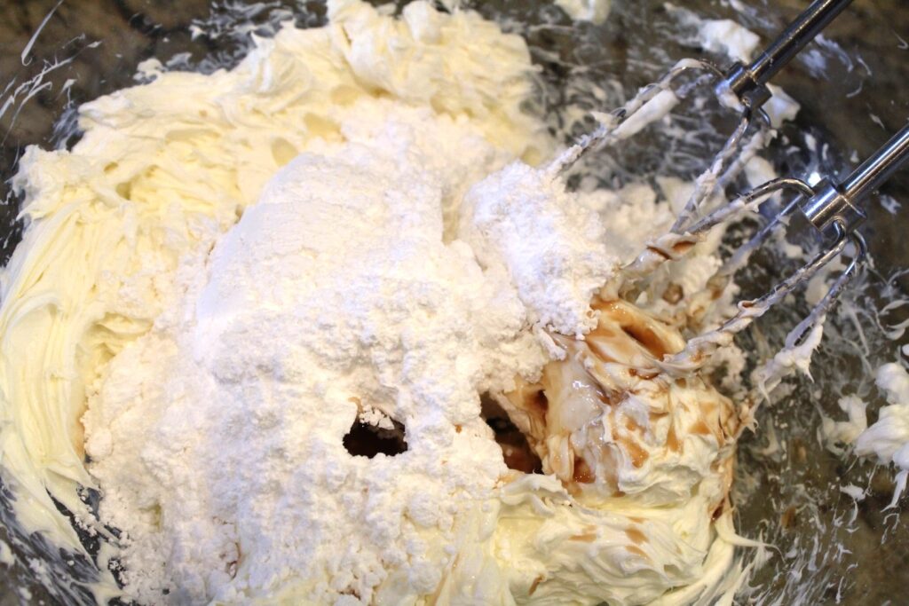 Powdered sugar and vanilla on top of whipped cream cheese in a glass bowl with hand mixer wands.