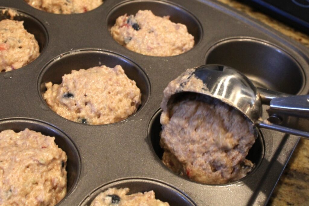 scooping banana berry muffin batter into a muffin tin with a large ice-cream scoop. 