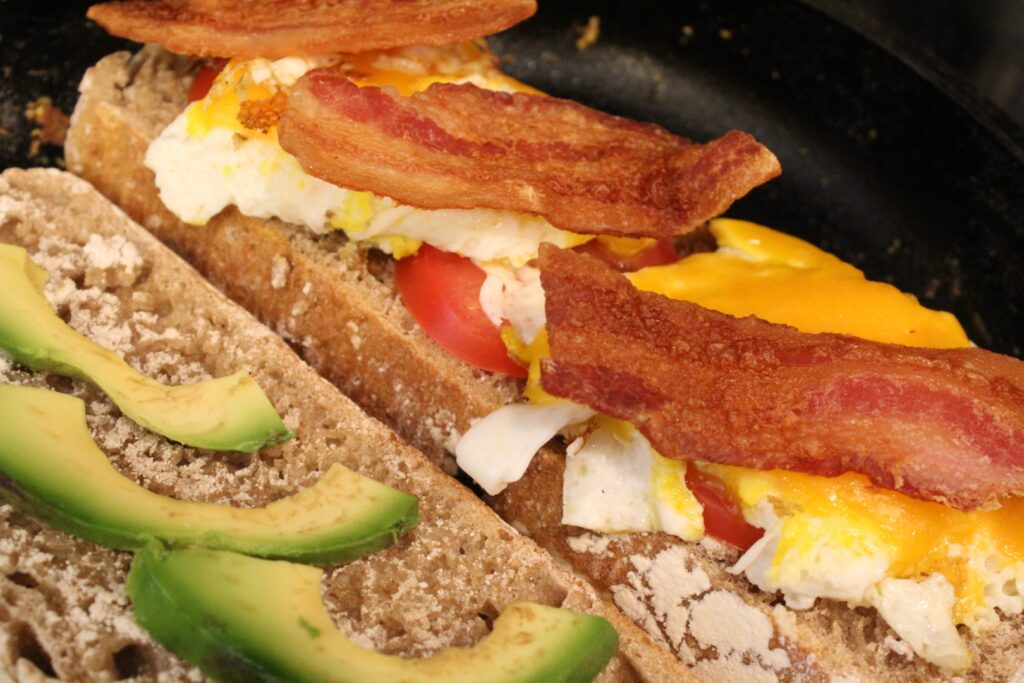 Adding avocado, eggs, tomatoes, and bacon to a toasting slice of sourdough bread. 