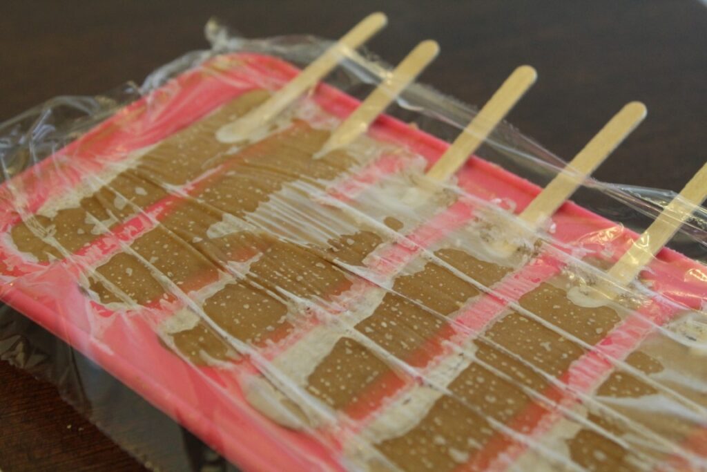 Frozen 2 ingredient peanut butter banana popsicles in a silicone ice stick tray covered in plastic wrap. 