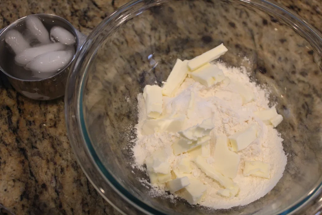 Butter cut into flour for a pie crust with a measuring cup full of ice water near by.