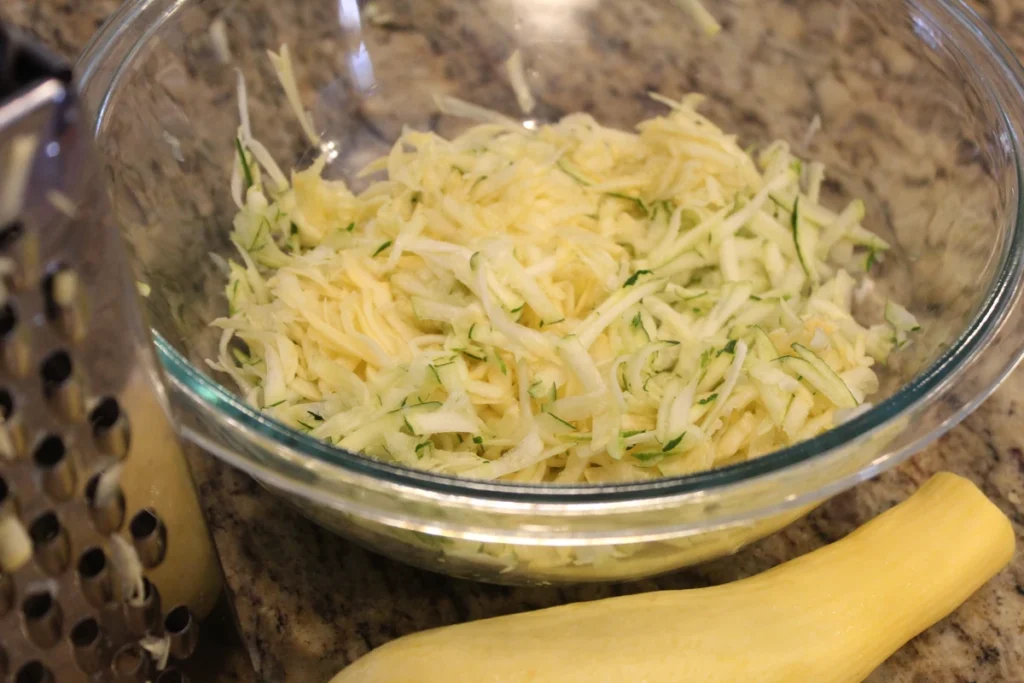bowl of shredded zucchini and yellow squash next to a grater and squash.