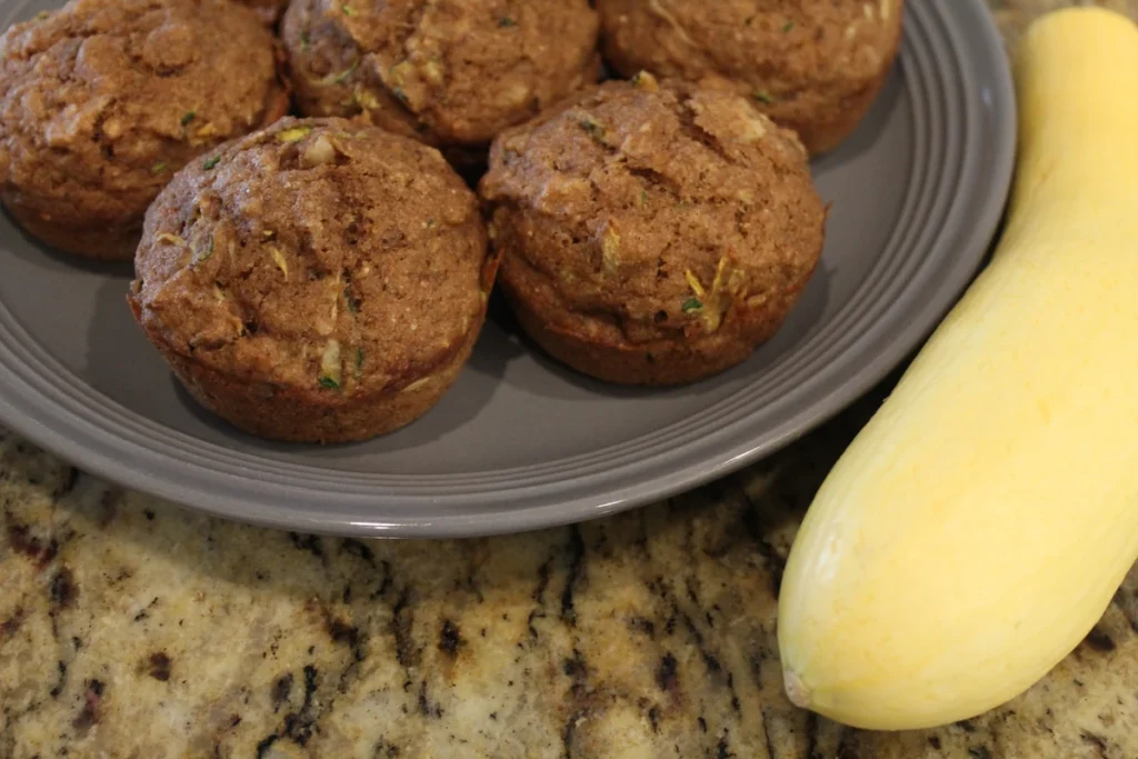 Plate of zucchini applesauce muffins beside a yellow squash.