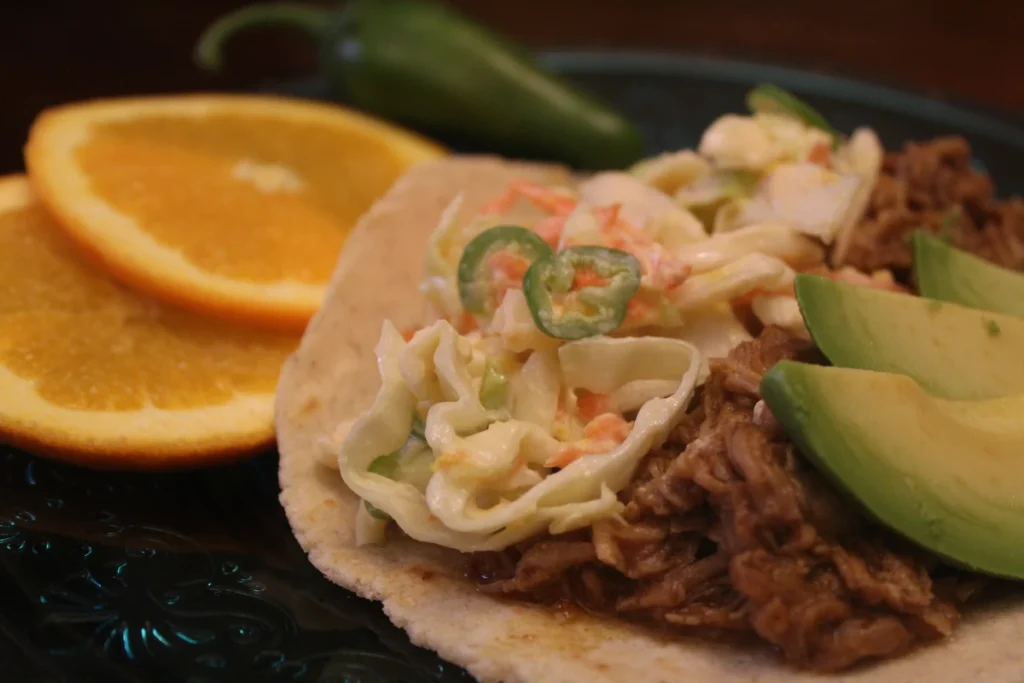 Close up of a citrus taco with sliced avocado and slaw, orange slices in the background. 