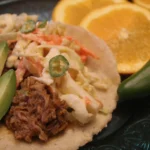 Orange pork taco topped with slaw, and sliced avocado with sliced oranges on the side.
