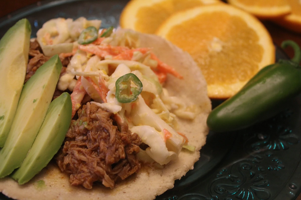 Orange pork taco topped with slaw, and sliced avocado with sliced oranges on the side.
